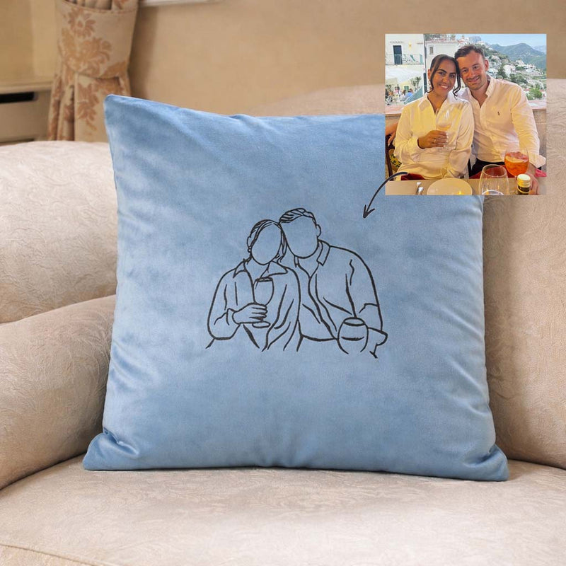 Blue pillow with a embroidered couple illustration on a beige couch, with a photo of a couple in the corner.