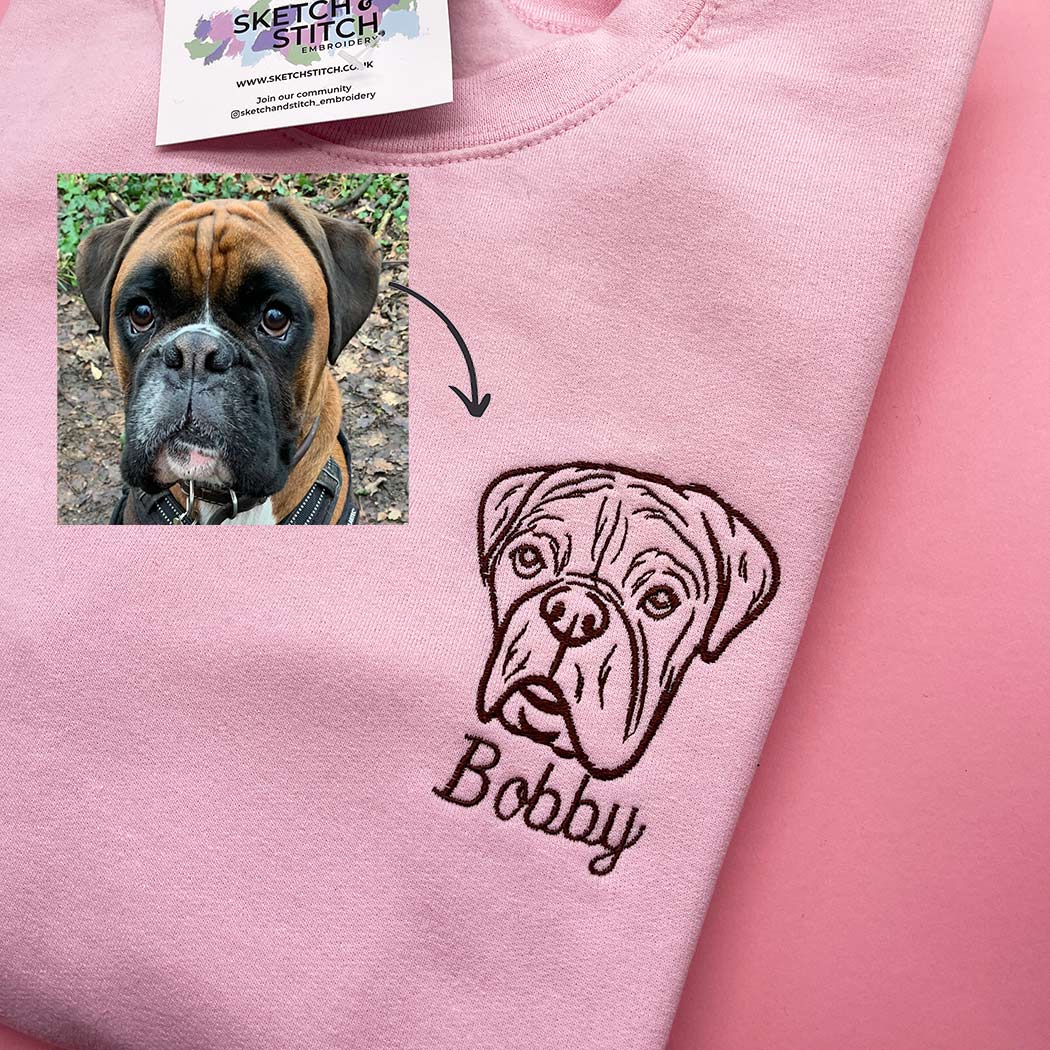 A ight pink sweatshirt on a pink background, featuring a black embroidered bulldog face and the name 'Bobby' on the chest. A small inset shows a real bulldog photo with an arrow pointing to the embroidery.