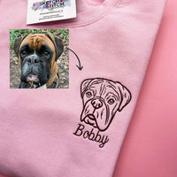 A ight pink sweatshirt on a pink background, featuring a black embroidered bulldog face and the name 'Bobby' on the chest. A small inset shows a real bulldog photo with an arrow pointing to the embroidery.