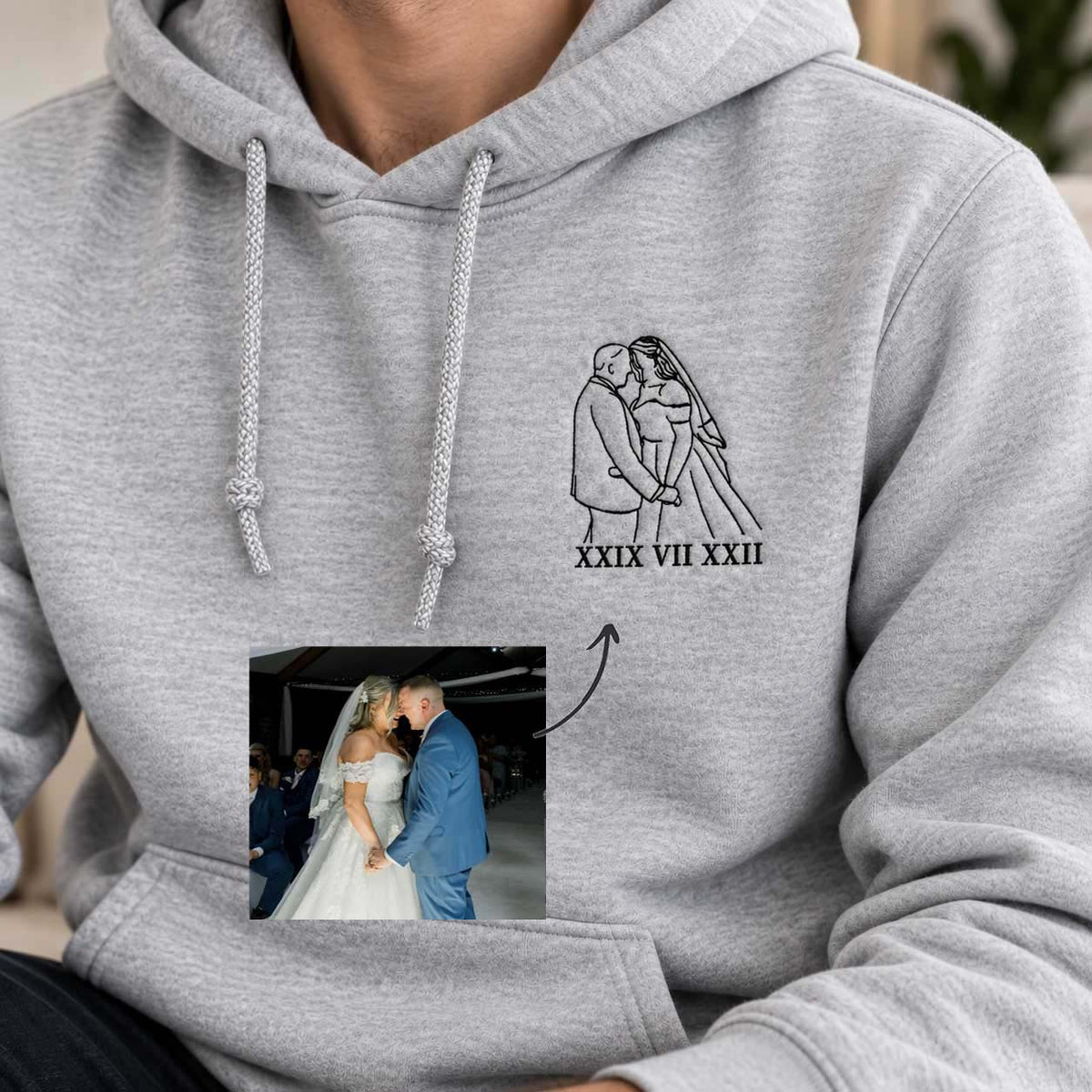 A close-up of a person wearing a light gray hoodie with a custom black line-art wedding graphic on the chest showing a couple and Roman numerals, plus an inset photo of the bride and groom.