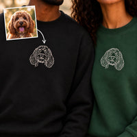 Two people wearing custom embroidered dog face designs on sweatshirts, one in black and one in dark green. An inset photo of a curly-haired dog appears in the top left with an arrow pointing to the embroidery.