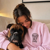 A woman in a light pink sweatshirt with a bulldog graphic and the name 'Bobby' stitched on it, hugging a brown and black Boxer dog on a beige couch.
