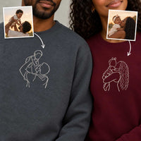 Two people wearing custom crewneck sweatshirts, one in dark gray and one in burgundy, both featuring a white line art graphic of a parent holding a child. Two inset photos of a father and child are shown with arrows pointing to the corresponding sweatshirt designs.