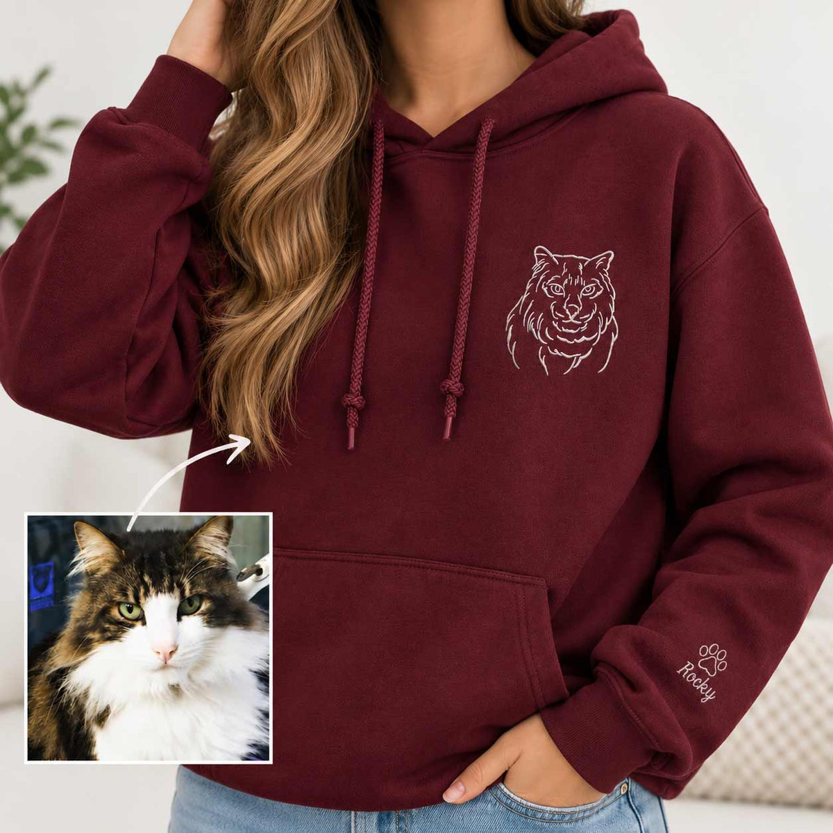 A woman wearing a burgundy pullover hoodie with a white tiger face graphic on the chest and a paw print with the name 'Rosy' on the sleeve. An inset photo on the bottom left shows a fluffy cat, which is linked to the custom name on the garment.
