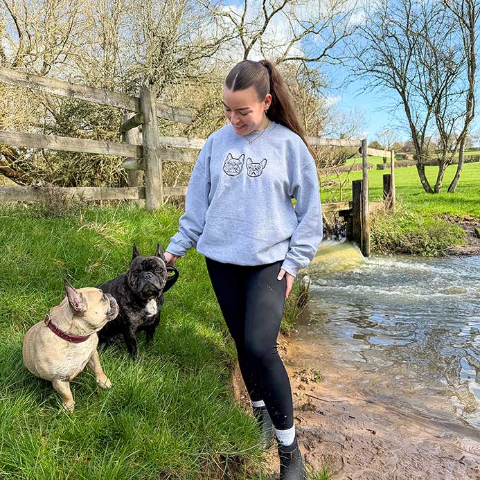 A woman stands outdoors by a small stream next to a wooden fence, wearing a light blue sweatshirt with a black cat graphic and black leggings, while interacting with two French bulldogs.