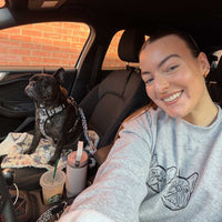 A woman smiling in a car next to a black French bulldog wearing a harness. The woman is wearing a light gray sweatshirt with a black dog graphic, and there are Starbucks cups on the car seat.