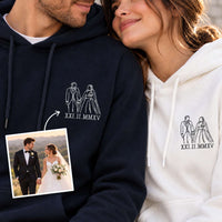 A man and a woman wearing matching custom hoodies, with the man on the left in a navy hoodie and the woman on the right in a white hoodie. Both hoodies feature a white line art graphic of a bride and groom holding hands above the Roman numerals 'XXI.