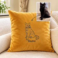 A mustard yellow throw pillow with a black line art drawing of a cat and the name 'Blackie' printed below, shown on a beige sofa. A small inset photo in the top right shows a real black cat, with an arrow pointing to the pillow design.