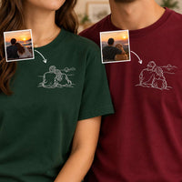 Two people wearing custom photo-printed t-shirts, one in forest green and one in burgundy, both featuring a white line art graphic of a couple sitting by the water. The shirts have been edited with inset photos of the couple and arrows pointing to the line art, demonstrating a custom photo-to-shirt print service.