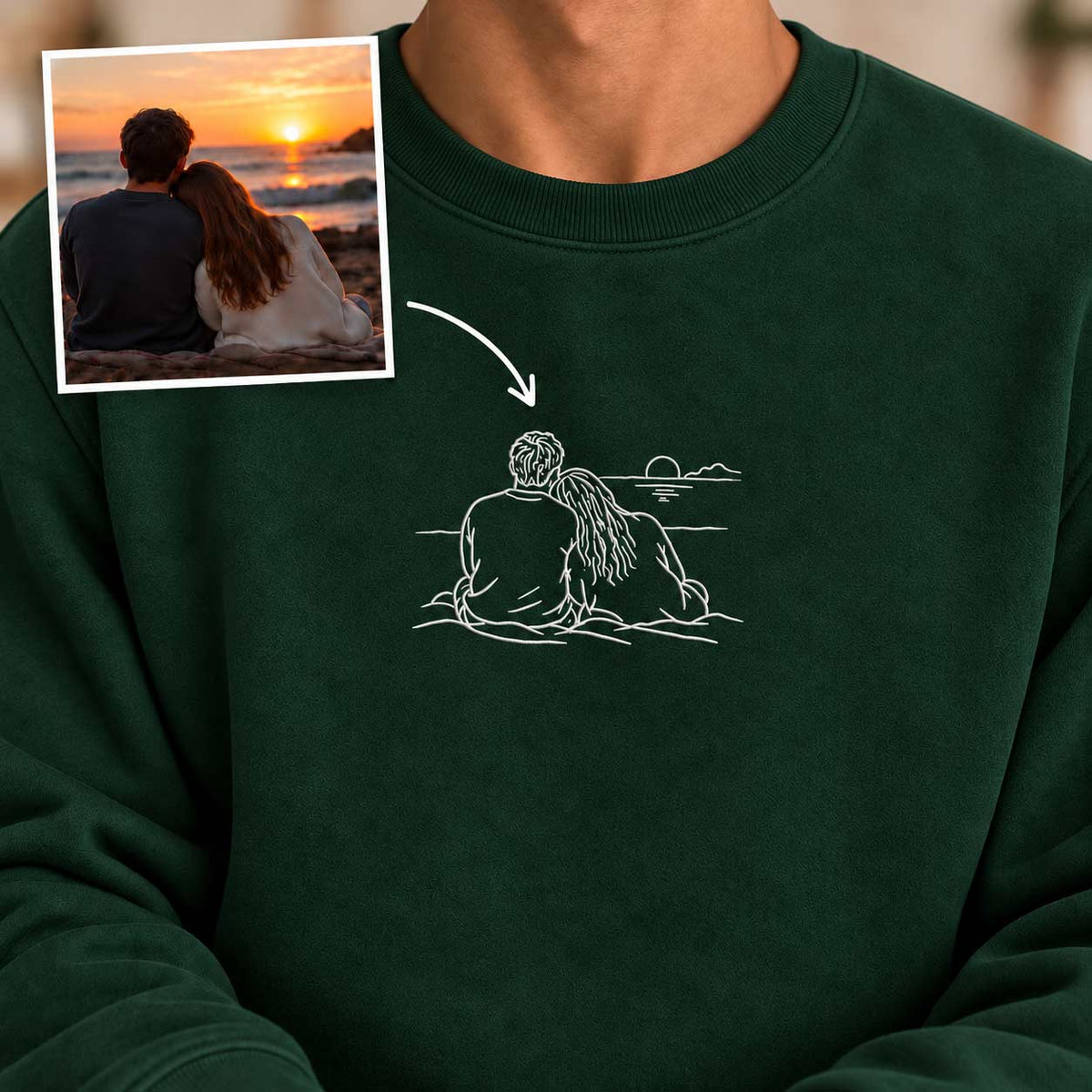 A dark forest green crewneck sweatshirt featuring a white minimalist line art graphic of a couple sitting by the ocean at sunset. A small inset image in the top-left corner shows the source photo used for the custom print, with an arrow pointing to the resulting design on the sweatshirt.