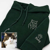 Dark forest green custom pet hoodie with white embroidered wolf outline and paw print with the name 'Rocky' on the sleeve. A small inset photo of a fluffy brown and white cat is shown in the lower-left corner with an arrow pointing to the hoodie's custom design area.
