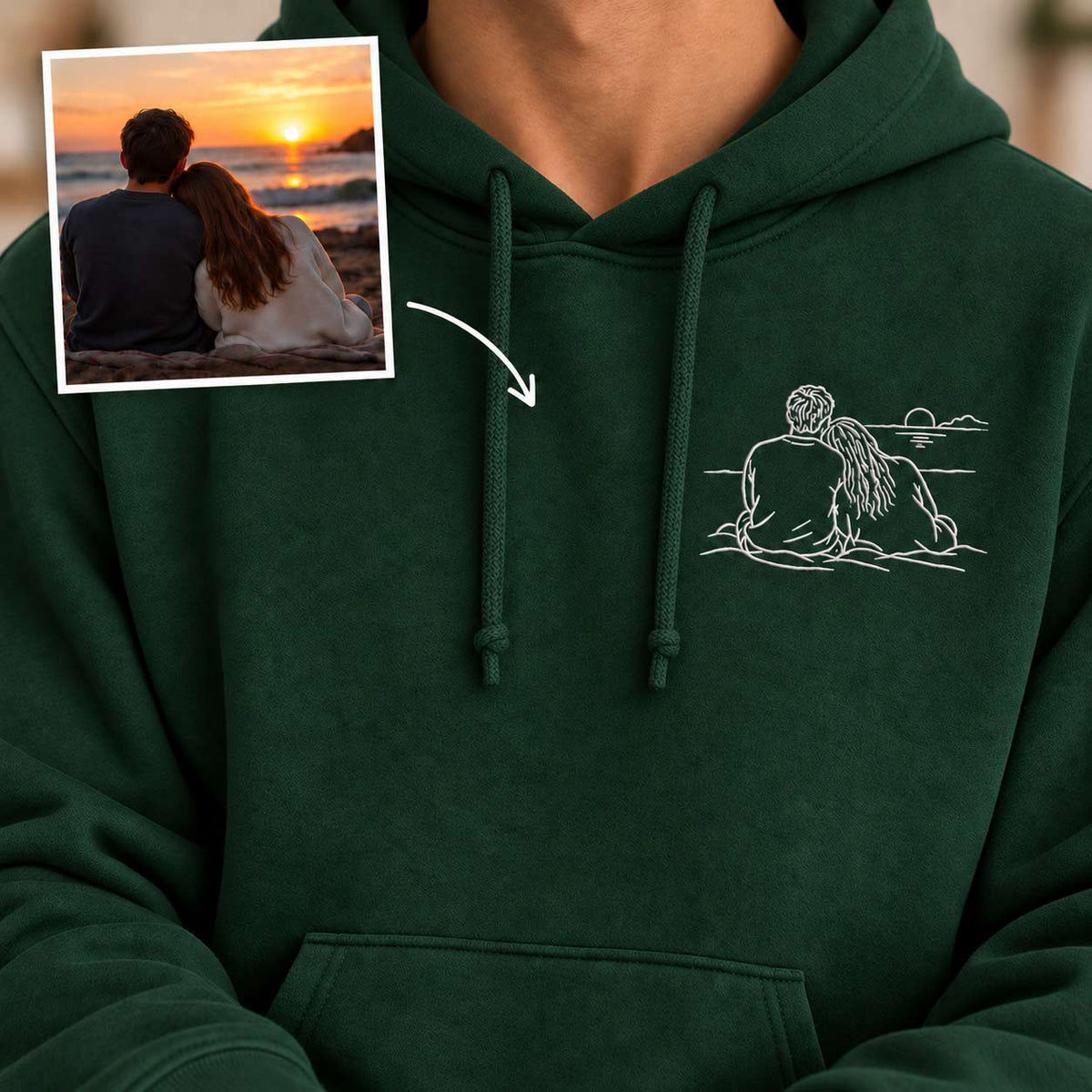 A dark green hoodie worn by a person, featuring a white line art graphic of a couple sitting by the ocean at sunset on the left chest. An inset photo in the top-left corner shows the couple by the beach at sunset, with an arrow pointing to the hoodie graphic.