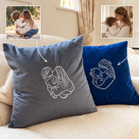 Two custom square throw pillows on a beige sofa, one in dark gray and one in royal blue, both printed with a white line drawing of a mother and child. Two small inset photos at the top show the source photo used for the custom print.