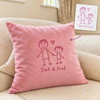 A pink throw pillow with a red hand-drawn stick figure family design and the text 'Dad & Fred' sits on a beige sofa. A small inset at the top right shows the original design sketch with a pointer arrow leading to the pillow.