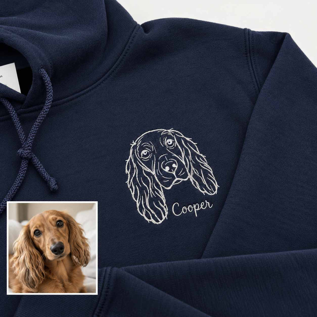A navy blue hoodie with a white embroidered line drawing of a dachshund and the name 'Cooper' on the chest. A small inset photo in the lower-left corner shows a real dachshund, likely used to demonstrate the custom pet design.