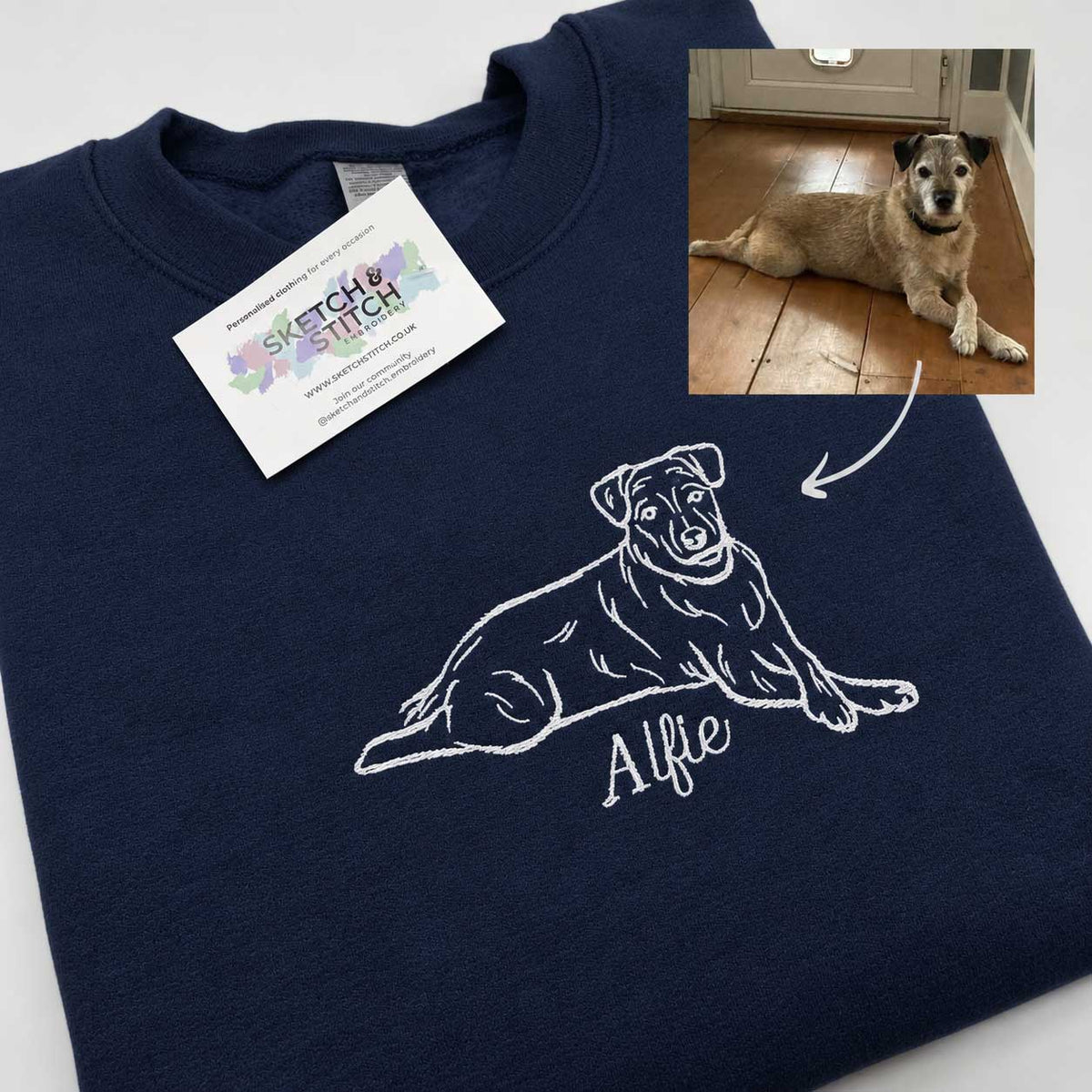 A navy blue crewneck sweatshirt laid flat on a white background, featuring a white line-art illustration of a dog and the name 'Alfie' printed on the front. A white brand tag reading 'Sketch & Stitch' is attached near the collar, and a small inset photo in the top-right shows a real dog lying on a wooden floor with an arrow pointing to the printed design.