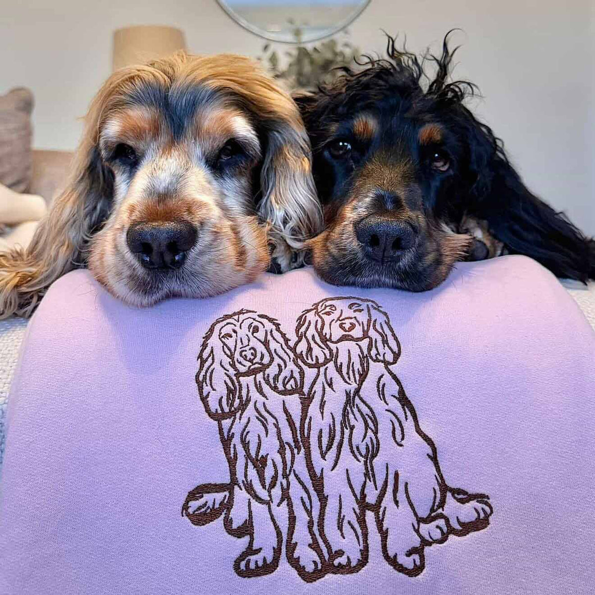 Two dogs resting on a lavender cushion with a brown embroidered design of two long-eared dogs, in a cozy indoor setting.