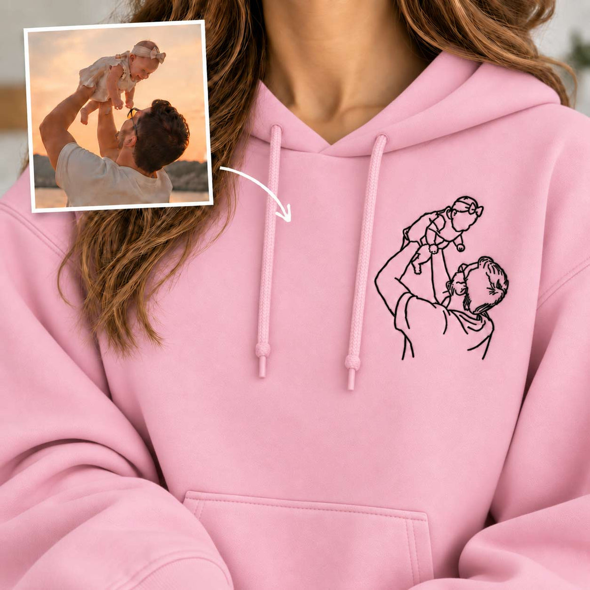 A close-up of a person wearing a light pink hoodie with a black line art graphic of a father holding a baby on the chest. An inset photo in the top-left corner shows a man holding a baby up outdoors at sunset, with an arrow pointing to the hoodie graphic, indicating a custom print-on-demand design.
