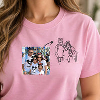 A close-up of a woman wearing a pink short-sleeve T-shirt. The shirt features a printed graphic on the chest showing a family photo on the left and a black line drawing of the same family on the right, connected by an arrow, likely representing a custom print-on-demand design service.