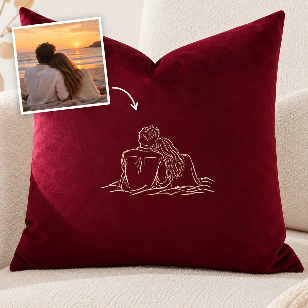 A deep burgundy velvet throw pillow with a white embroidered line drawing of a couple sitting together, shown on a beige sofa. A small inset photo in the top-left corner shows a couple by a sunset, with an arrow pointing to the pillow embroidery, indicating a custom design service.
