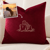 A deep burgundy velvet throw pillow with a white embroidered line drawing of a couple sitting together, shown on a beige sofa. A small inset photo in the top-left corner shows a couple by a sunset, with an arrow pointing to the pillow embroidery, indicating a custom design service.