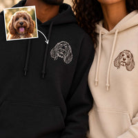 Two people wearing custom pet hoodie prints, with a small inset photo of the dog used for the embroidery design. One hoodie is black with a white dog portrait, and the other is cream with a brown dog portrait.