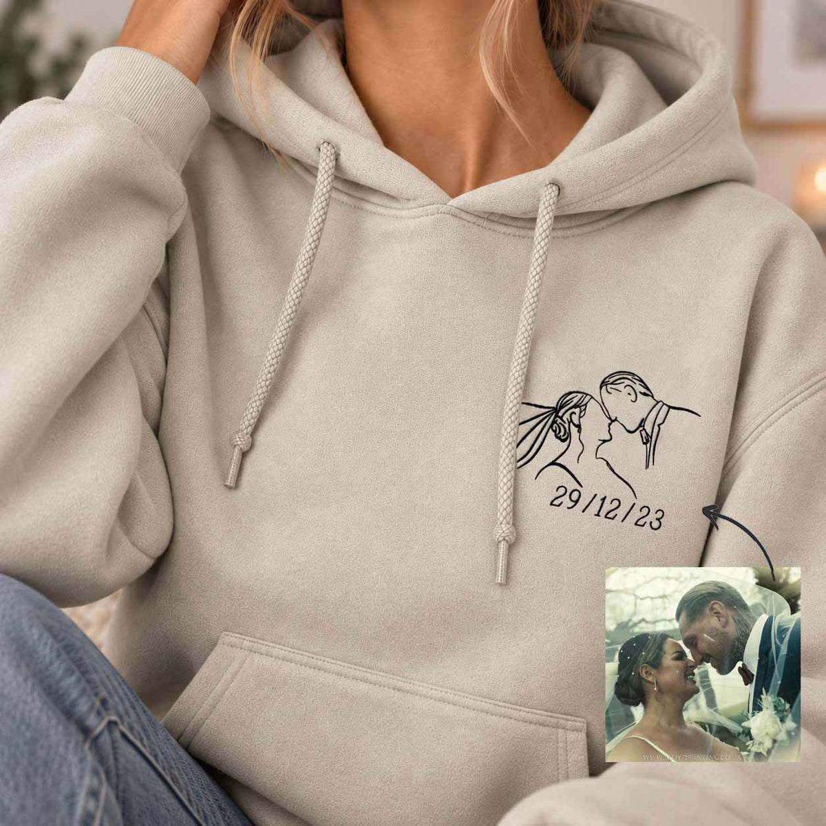 A person wearing a light beige hoodie with a black line-art couple illustration and the date '29/12/23' on the left chest. A small inset photo in the lower right shows the couple, likely used as a customization example.