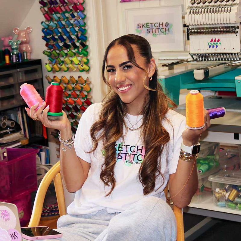 A woman sitting in a chair in a craft room, holding two colorful spools of thread, one pink with a green base and one orange with a purple base.