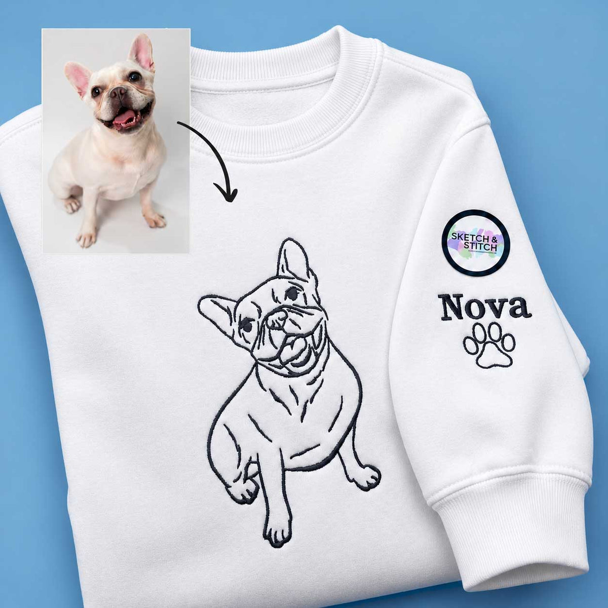 A white crewneck sweatshirt laid flat on a blue background, featuring a black line-art illustration of a French bulldog on the chest and a custom sleeve print with a paw print, the name 'Nova', and a circular 'Special Stitch Studio' logo. A small inset photo of a real French bulldog with an arrow points to the drawing, indicating a custom pet illustration service.