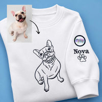 A white crewneck sweatshirt laid flat on a blue background, featuring a black line-art illustration of a French bulldog on the chest and a custom sleeve print with a paw print, the name 'Nova', and a circular 'Special Stitch Studio' logo. A small inset photo of a real French bulldog with an arrow points to the drawing, indicating a custom pet illustration service.